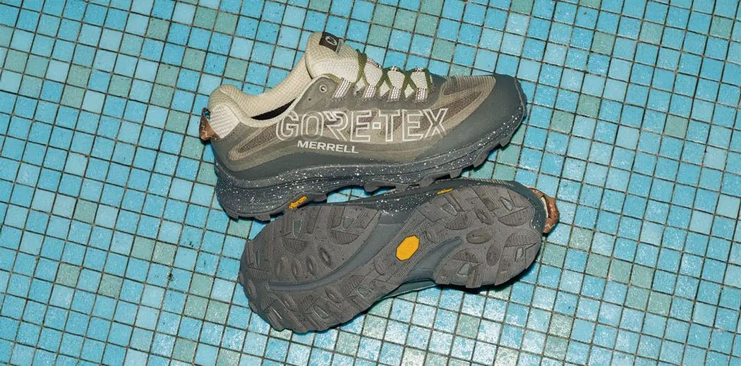 Gray Gore-Tex Merrell hiking shoes on a blue tile background. One shoe is side view; the other shows the rugged sole. Outdoorsy and adventurous vibe.