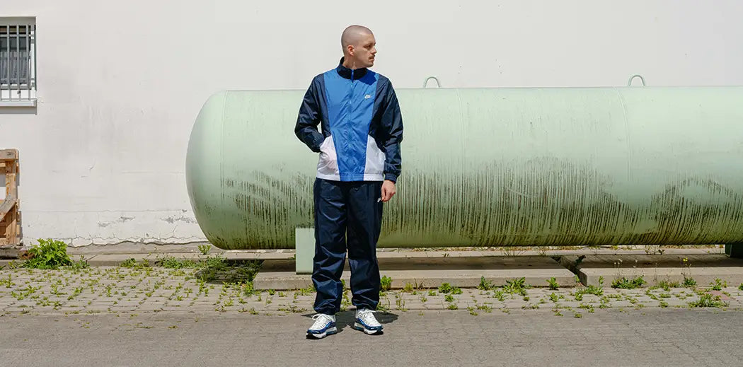 A person in a blue and white tracksuit from Nike with Nike Air Max 98 on feet stands on a paved surface in front of a large, cylindrical tank and a white building. The mood is calm and relaxed.