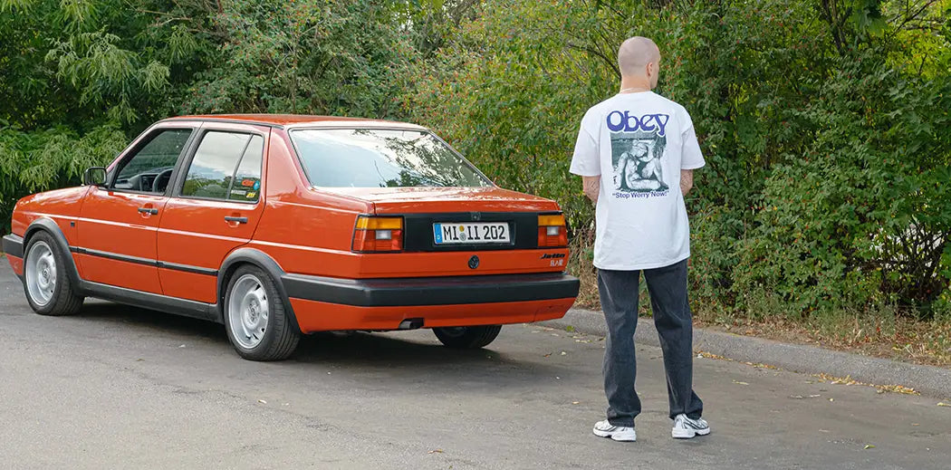 A person in a white graphic T-shirt from Obey and baggy denim jeans stands on a quiet road beside a red vintage car. The backdrop is lush green trees, conveying a tranquil atmosphere.