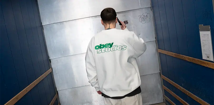 A person in a white "Obey Studios" sweatshirt stands in an industrial-looking hallway with blue walls and metal doors, holding a phone to their ear.