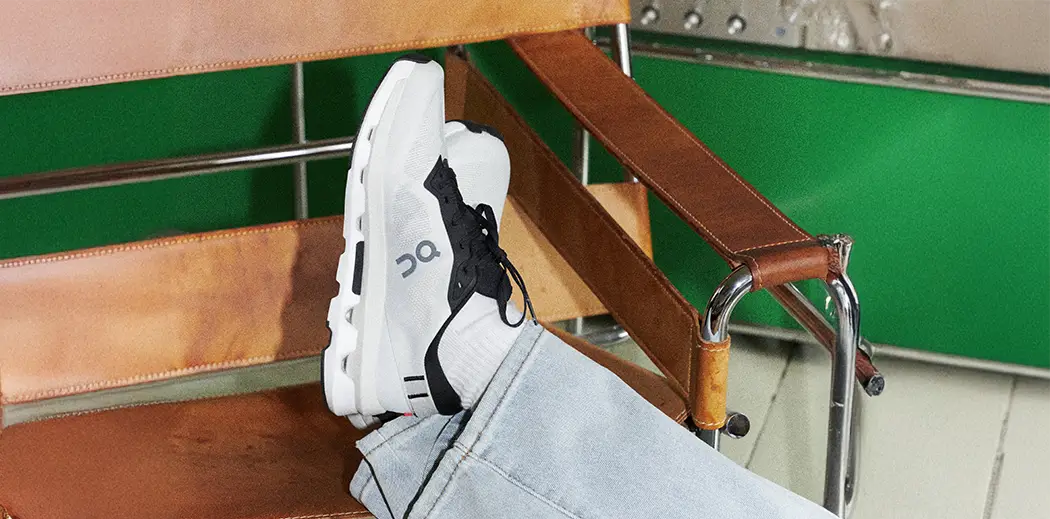 Casual scene with legs wearing light jeans and white sneakers from On Running Cloud Technology resting on a tan leather chair. Background shows green decor and stereo equipment.