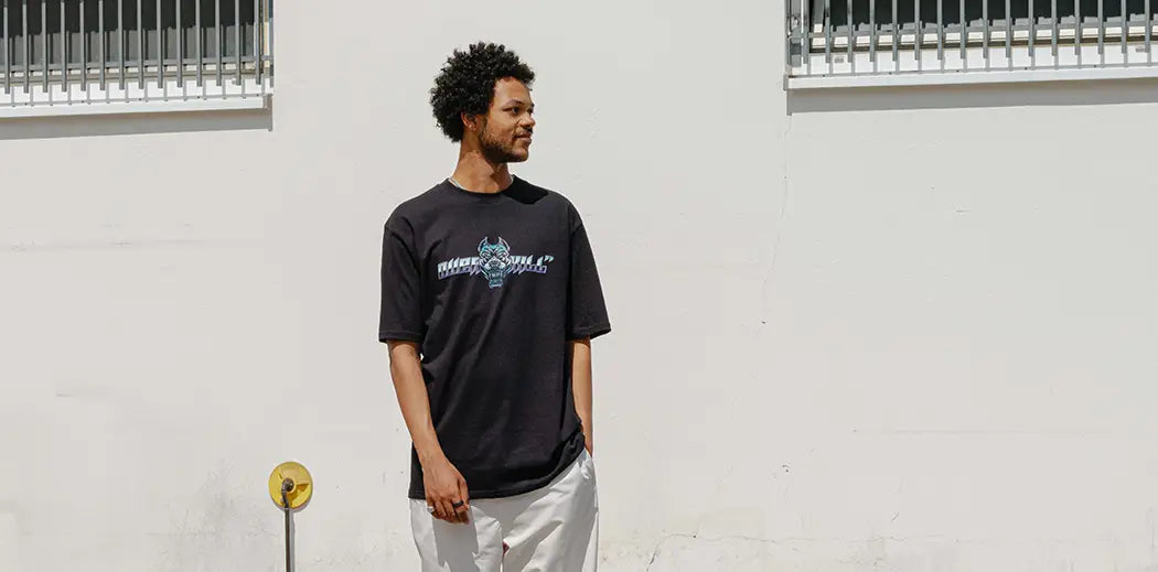 Man in casual attire stands in an urban setting. He wears a black T-shirt with a bulldog made my famous sneaker store from Berlin OVERKILL and light pants, against a white wall with barred windows, under a clear sky.