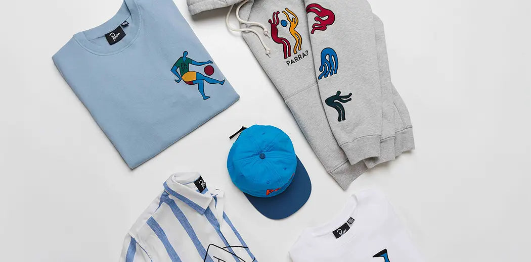 Assorted stylish clothing from streetwear brand PARRA, items are laid out, including a light blue tee with playful graphics, a gray hoodie with colorful abstract designs, a striped shirt, and a blue cap.