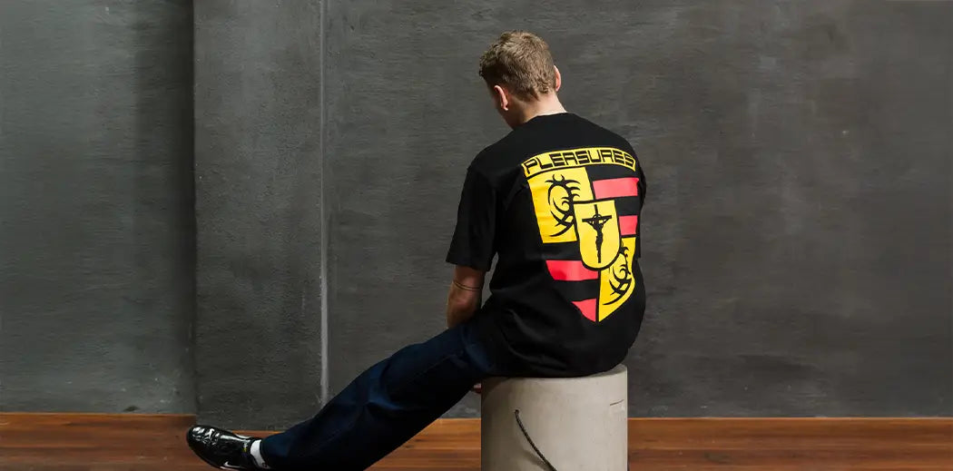 A person in a black streetwear brand PLEASURES t-shirt with a bold yellow and red shield design sits on a concrete stool, facing a textured dark gray wall, on a wooden floor.