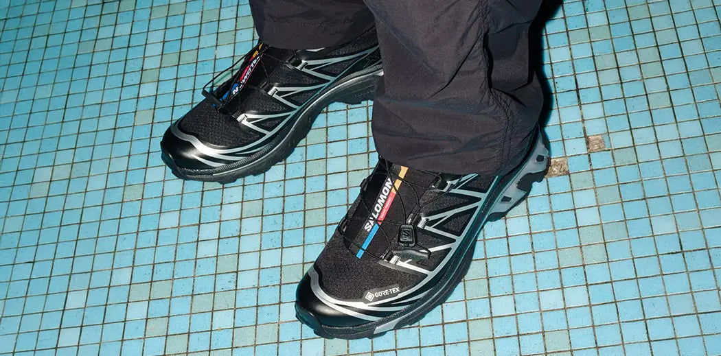 Person wearing black sneakers from Salomon with silver accents and GORE-TEX label, standing on a blue tiled floor. The mood is casual and sporty.