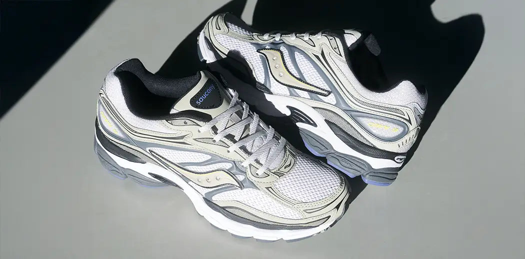 A pair of stylish gray and white Y2K Saucony sneakers with black lining and intricate patterns, casting a shadow on a smooth gray background, conveying sleekness.