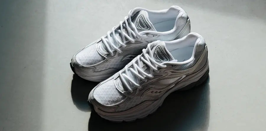 A pair of stylish gray and white Y2K Saucony sneakers with black lining and intricate patterns, casting a shadow on a smooth gray background, conveying sleekness.
