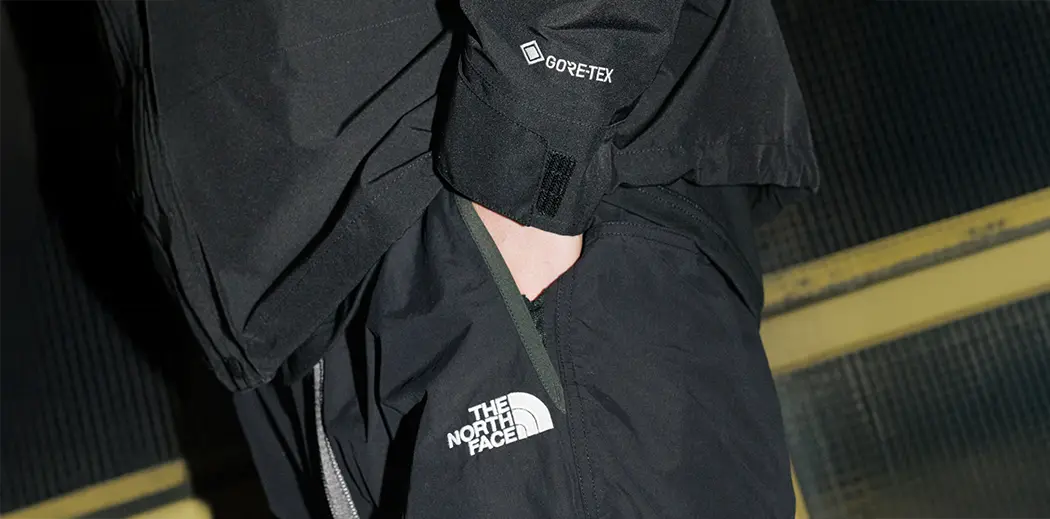 Person wearing black Gore-Tex The North Face clothing. The shot focuses on logos, emphasizing weatherproof attire against a dark, urban background.