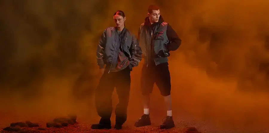 Two individuals pose confidently in gray bomber jackets from Alpha Industries and dark pants amid thick orange smoke, evoking a mysterious, edgy atmosphere.