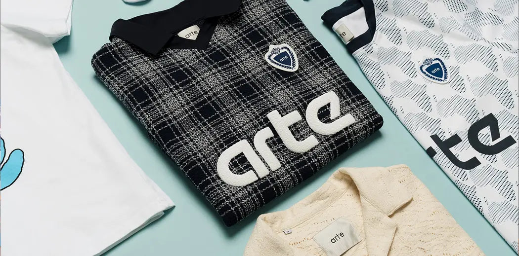 Folded Arte Antwerp Streetwear clothing items on a light blue background: white and patterned shirts, socks, cream knit, and jeans with "Arte" logo, creating a modern, stylish vibe.