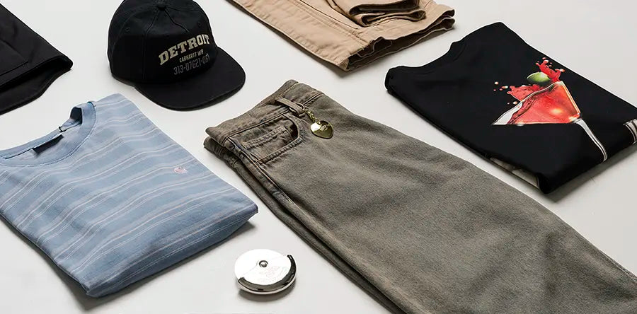 Folded casual Carhartt WIP clothing and accessories on a white surface, including a striped shirt, black cap, jeans with keychain, and a graphic tee. Stylish and laid-back vibe.