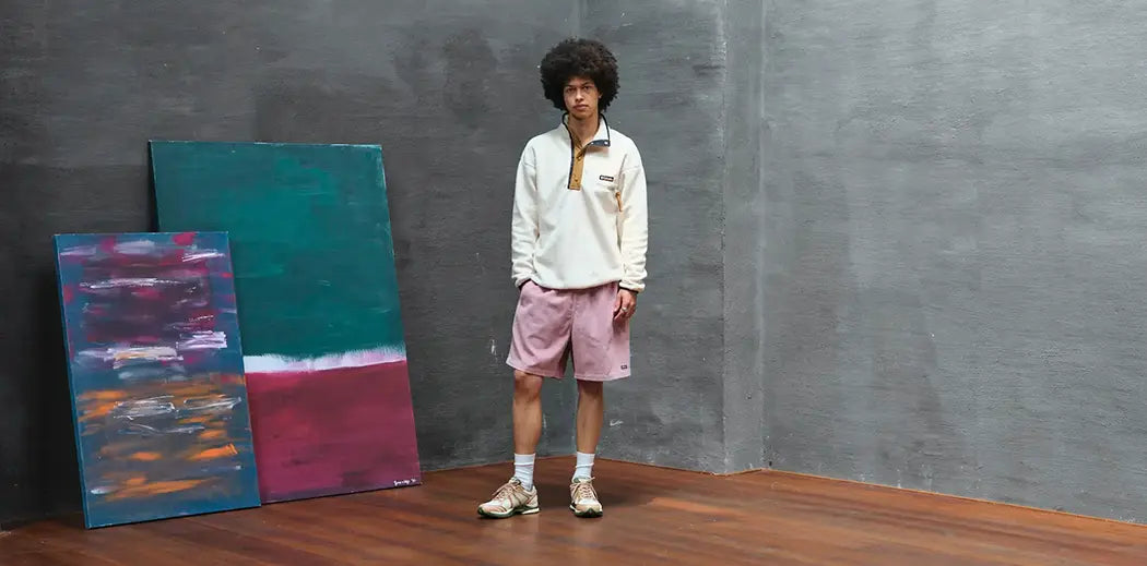 A person in casual attire and a Columbia Fleece Sweatshirt stands in a minimalist room with wooden flooring, beside two abstract paintings with green, blue, pink, and orange tones.