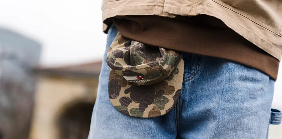 A person wearing a tan jacket and blue jeans, with a camo-patterned cap from Columbia tucked into the waist. The background is blurred, suggesting an outdoor setting.