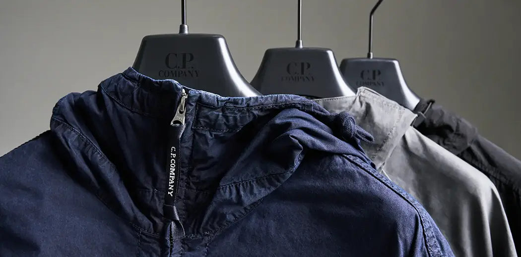 Three C.P. Company jackets in navy, gray, and black hang on branded hangers. The focus is on their textured fabric and sleek, modern design.