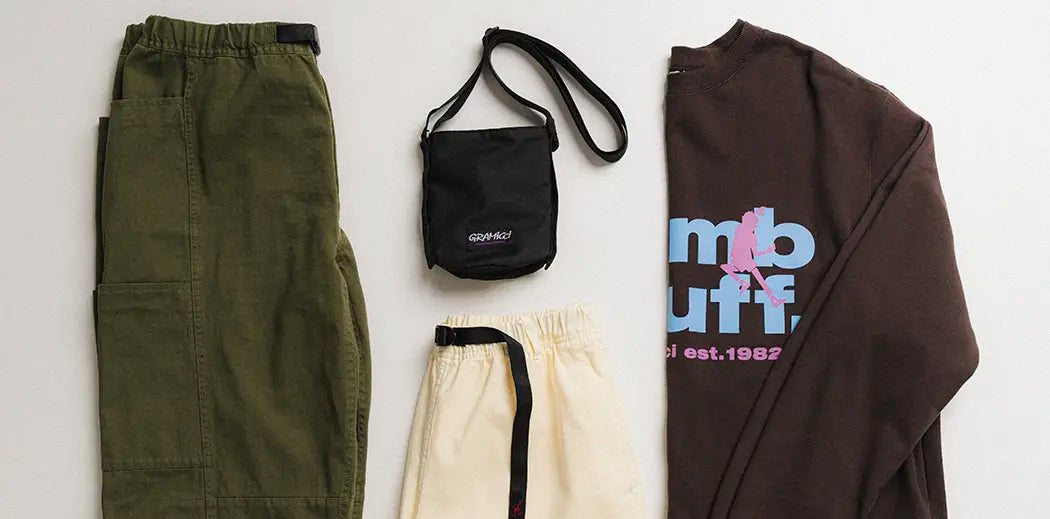 Four clothing items from Streetwear Outdoor brand Gramicci are neatly arranged: olive green pants, cream shorts, a brown graphic sweatshirt, and a small black crossbody bag. Casual style.