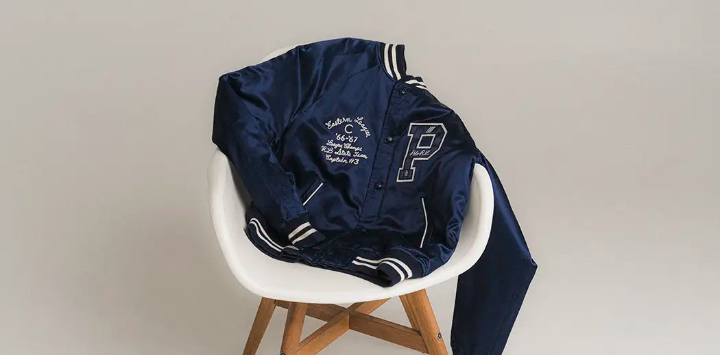A navy blue varsity jacket from Polo Ralph Lauren with white striped cuffs and collar draped over a white chair. The jacket features a large letter "P" and embroidered text.