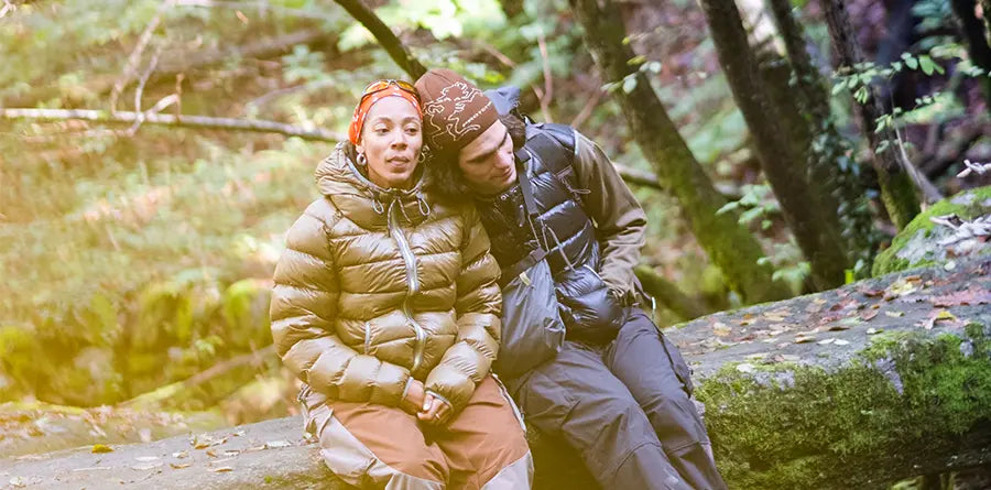 A couple in warm jackets of Ka_Yo_Prototype sits on a mossy log in a forest. The man leans his head on the woman's shoulder, conveying tranquility and closeness.