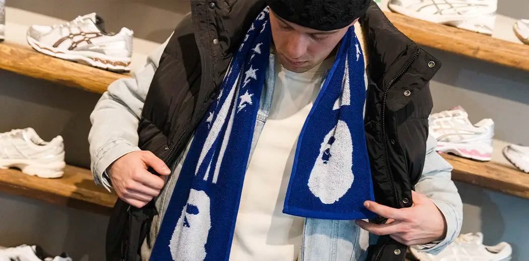A person adjusts their black puffer vest over a light denim jacket, wearing a blue scarf from BAPE with white patterns. Background: shelves with sneakers.
