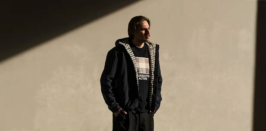A man in a black jacket and graphic T-shirt stands against a sunlit beige wall, gazing to the side as sharp shadows create a calm, contemplative mood.