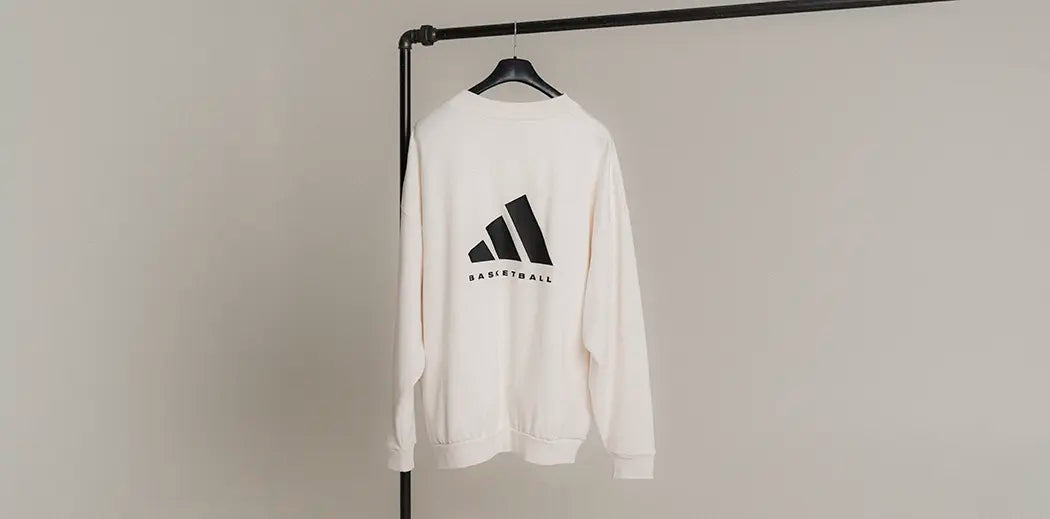 A white sweatshirt from adidas Basketball designed by Jerry Lorenzo from Fear of God hangs on a clothing rack. It features a black logo and the word "Basketball" on the back. The setting is minimalistic and neutral-toned.