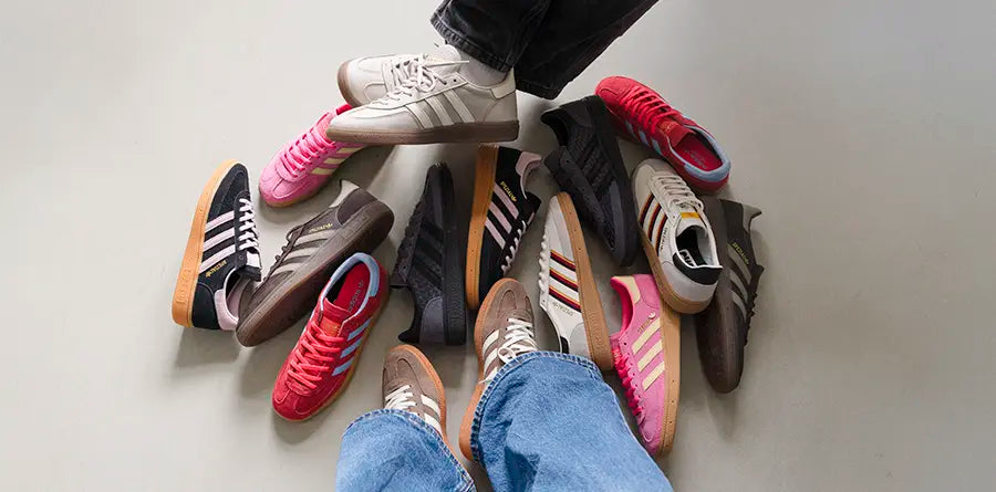 Two pairs of feet showcase various adidas Handball Spezial sneakers in different colors, like pink and red, surrounded by ten scattered shoes on a light floor.