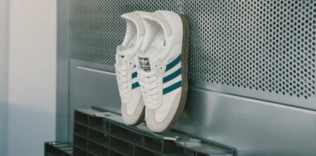 A pair of white adidas Samba sneakers with blue stripes, with gum sole leaned against a perforated metal surface. The style is classic and minimalistic.