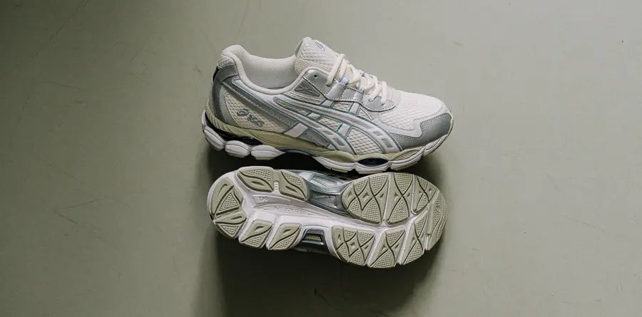 Two white and gray Asics Gel-Nyc sneakers are placed on a plain surface. One shoe is upright, showing the side with a logo, while the other reveals its textured sole.