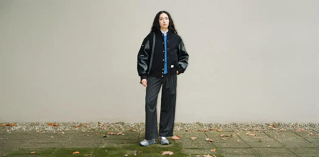 Person with long dark hair wearing a black varsity jacket or college jacket, blue shirt, striped pants, and sneakers. Stands on mossy pavement with fallen leaves.