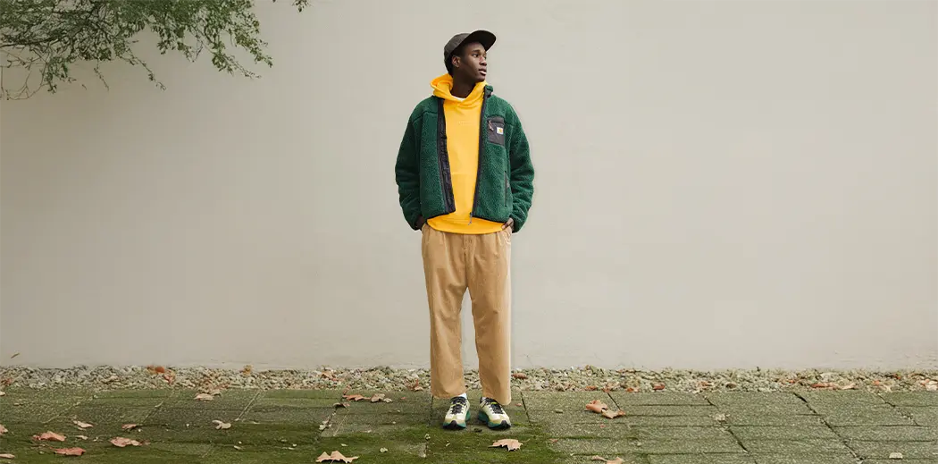 A person stands outdoors against a plain wall, wearing a green fleece jacket, yellow hoodie, tan pants, and sneakers. Fallen leaves scatter the ground.
