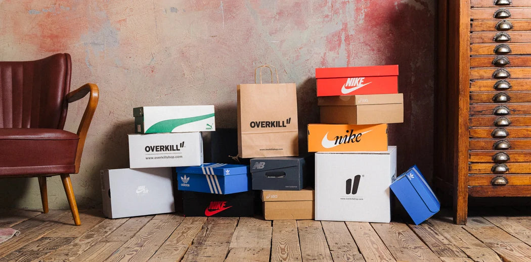 A stack of branded shoe boxes, including Nike, Adidas, and Overkill, arranged on a wooden floor beside a vintage chair and cabinet, against a textured wall.