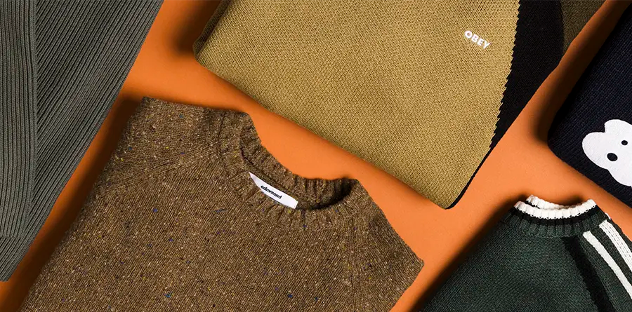 Cozy knitwear sweaters displayed on an orange surface, featuring textures and patterns. Includes muted green and brown hues, with one sweater showing "OBEY."