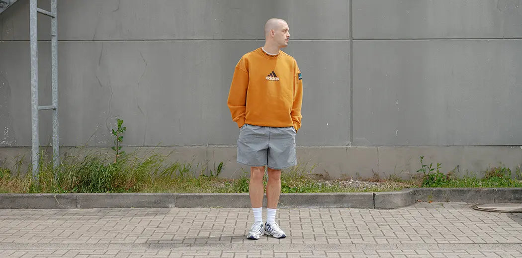 A person in an orange adidas Equipment sweater and gray shorts and y2k adidas running sneakers stands on a paved area, gazing sideways. The backdrop is a gray wall with a metal staircase.