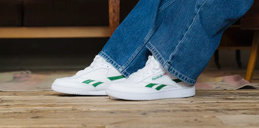 A person lacing up their Reebok Club C sneakers on a wooden floor.