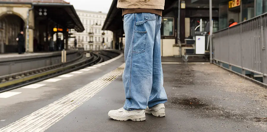 Man in baggy jeans and sneakers stands on a wet train platform, gazing down curved tracks. Overcast weather sets a reflective tone.
