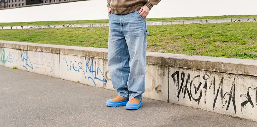 Person in loose blue jeans and brown shoes with blue soles stands on a graffiti-covered ledge next to green grass, creating a casual urban vibe.
