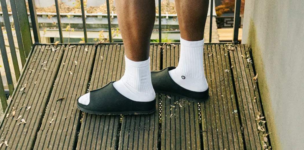 Person wearing black slide sandals with white socks stands on a wooden deck. The setting is outdoors with scattered leaves, conveying a casual vibe.