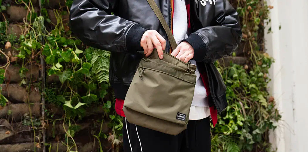 Person in a black jacket holding an olive green crossbody bag, standing in front of a lush green vertical garden, exuding a casual, urban vibe.