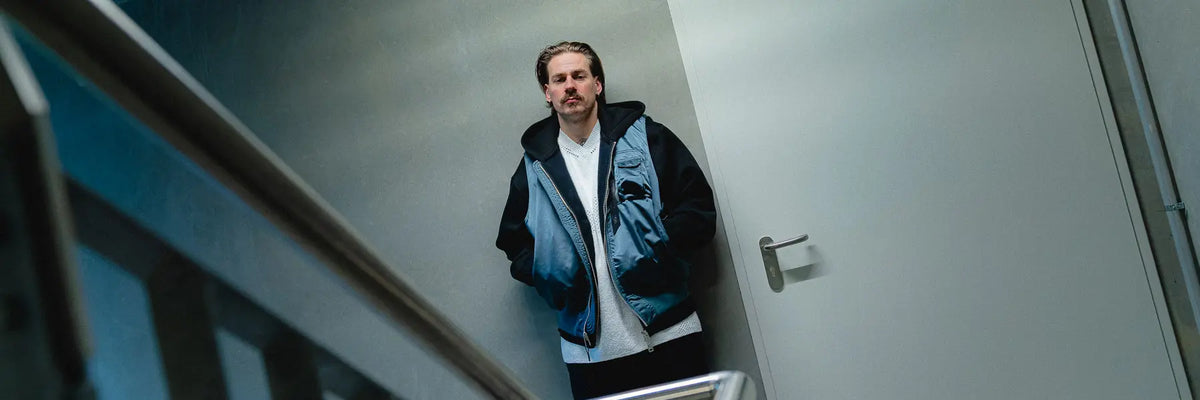 Man wearing a blue Alpha Industries vest over a black hoodie standing in a narrow hallway near a closed door