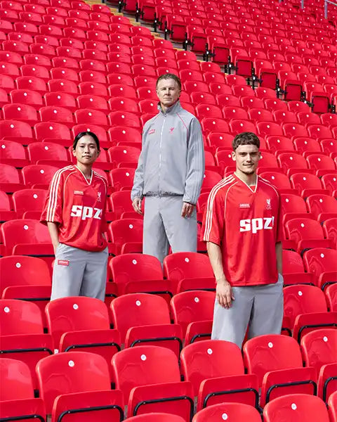Two people in red sports jerseys and gray pants and one person in a gray tracksuit standing among red stadium seats