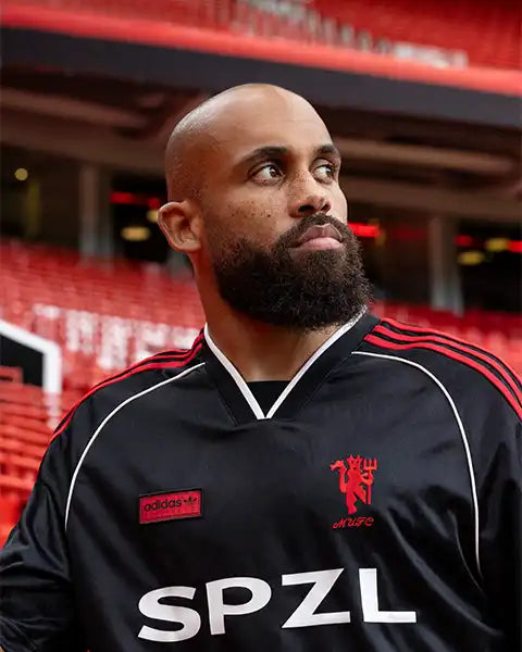 Bearded man wearing a black Adidas SPZL sports jersey looking to the side in a stadium