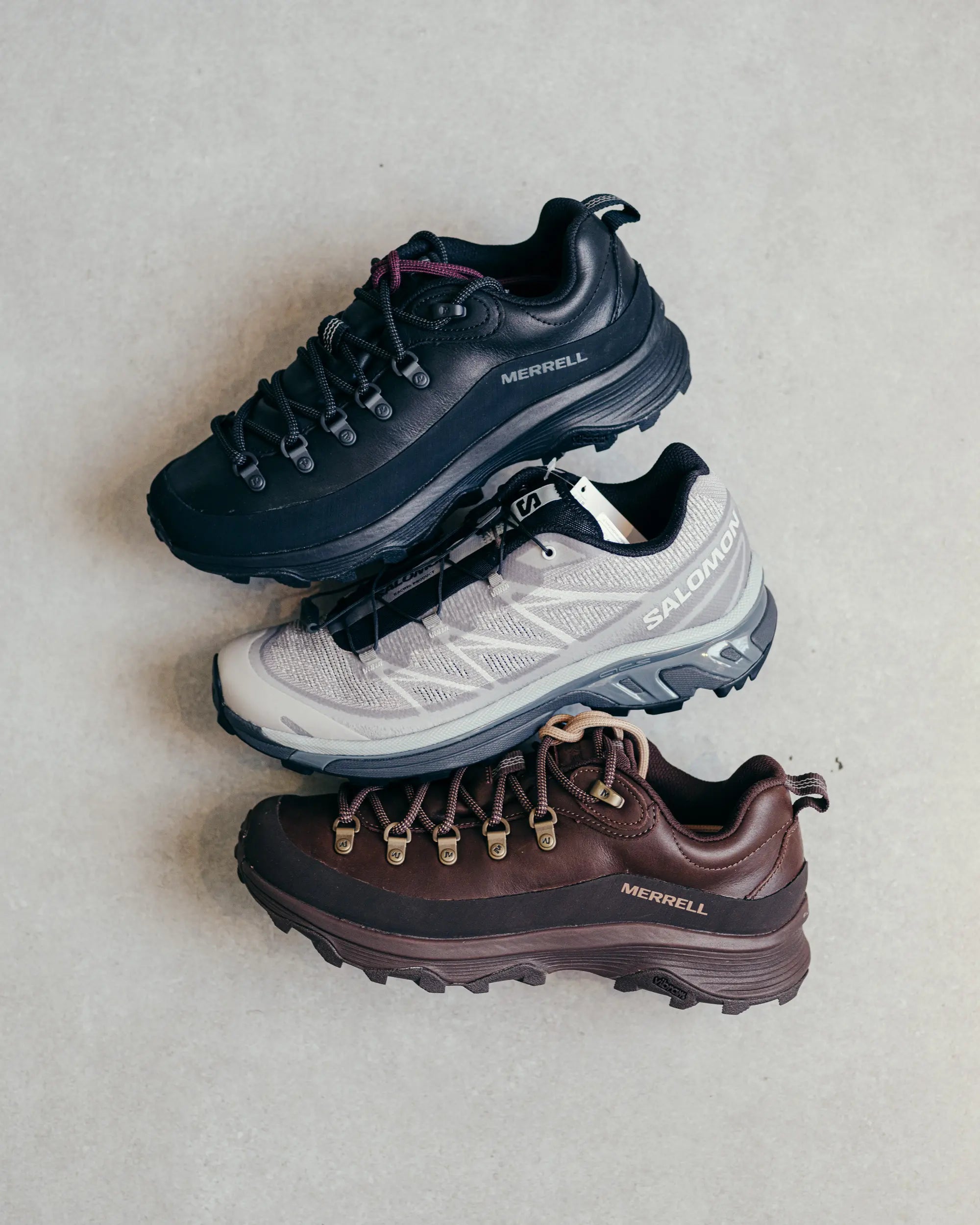 Three hiking shoes from Merrell and Salomon arranged vertically on a light surface. Top is black, middle is gray with white, and bottom is brown. Each shoe has visible brand logos.