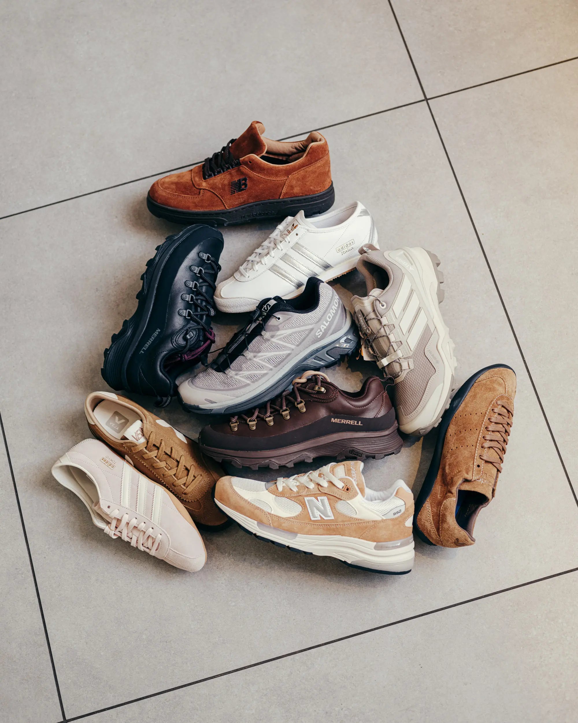 A circle of various sneakers on a tiled floor. The shoes, in earthy tones like brown, beige, and white, include both athletic and casual styles.
