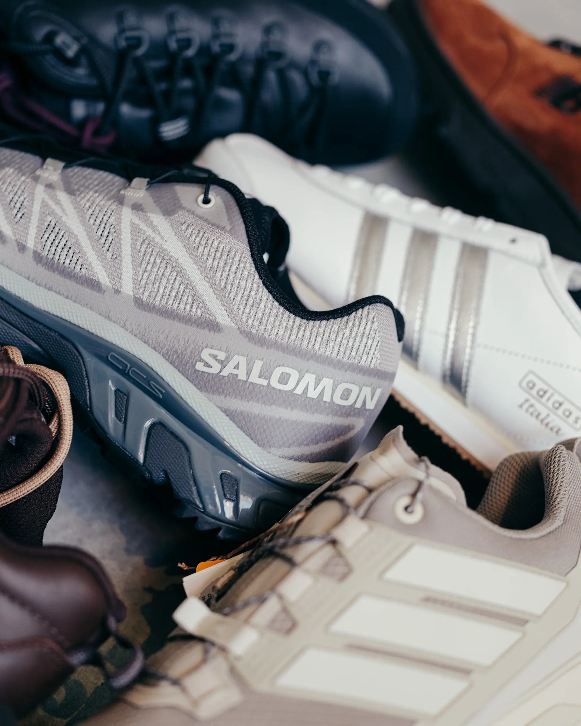 A close-up of various sneakers, featuring a gray Salomon shoe prominently. White and brown shoes surround it, creating a casual, sporty vibe.