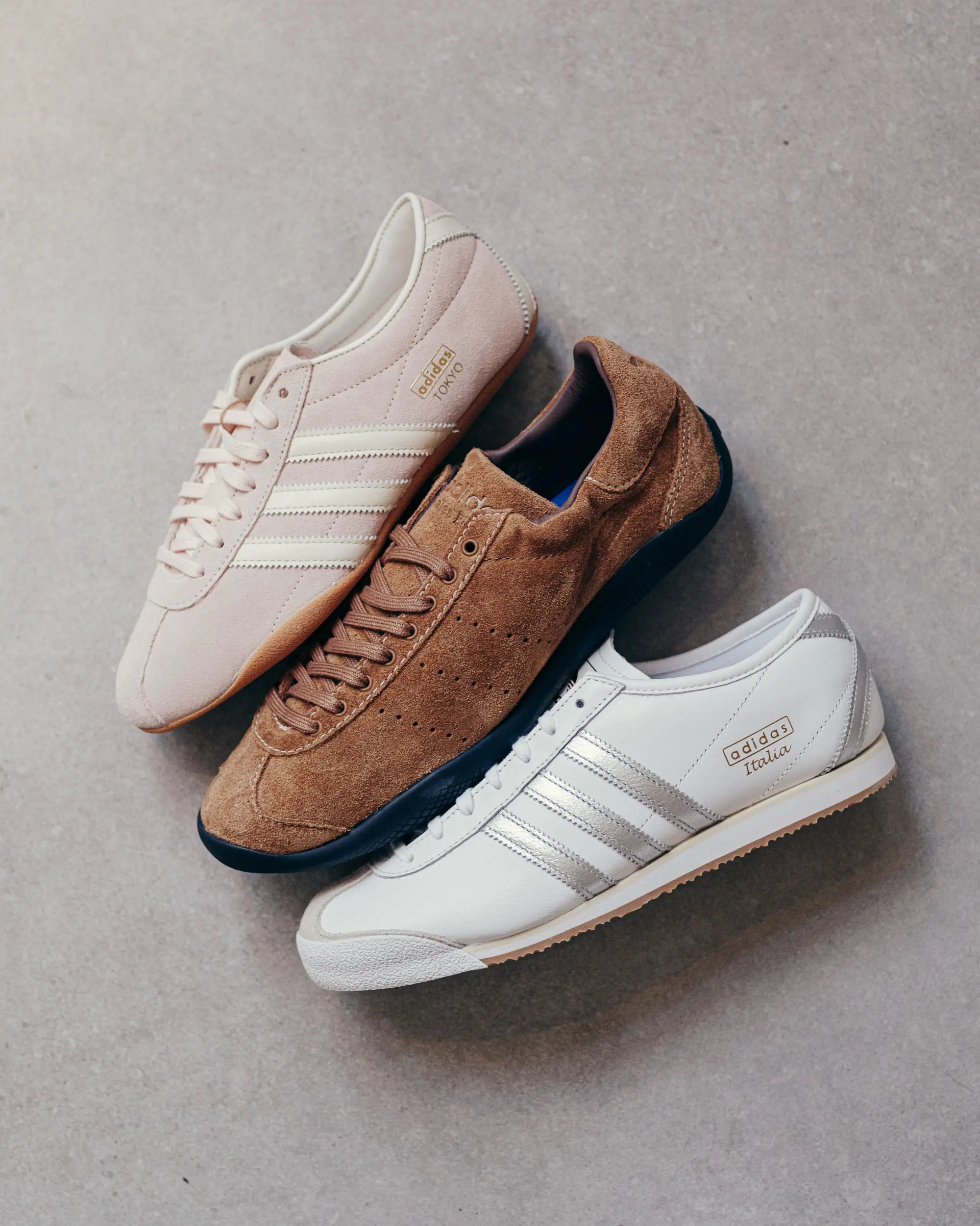 Three Adidas shoes are arranged on a gray surface. A beige shoe is on top, a brown suede shoe in the middle, and a white shoe with silver stripes at the bottom.