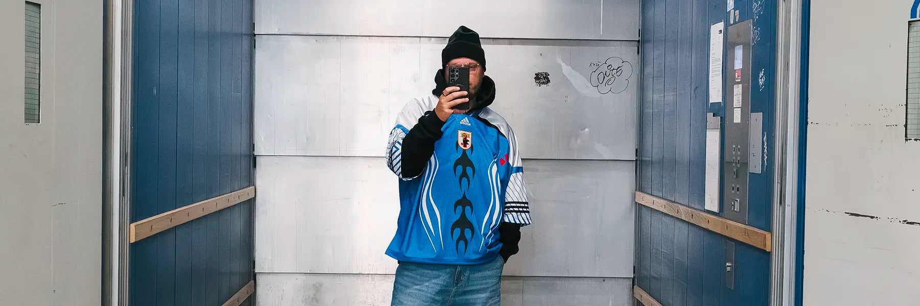 A person in a blue Japan jersey and jeans takes a selfie in an industrial elevator. The mood is casual, and the background is metallic with graffiti. Desktop Banner
