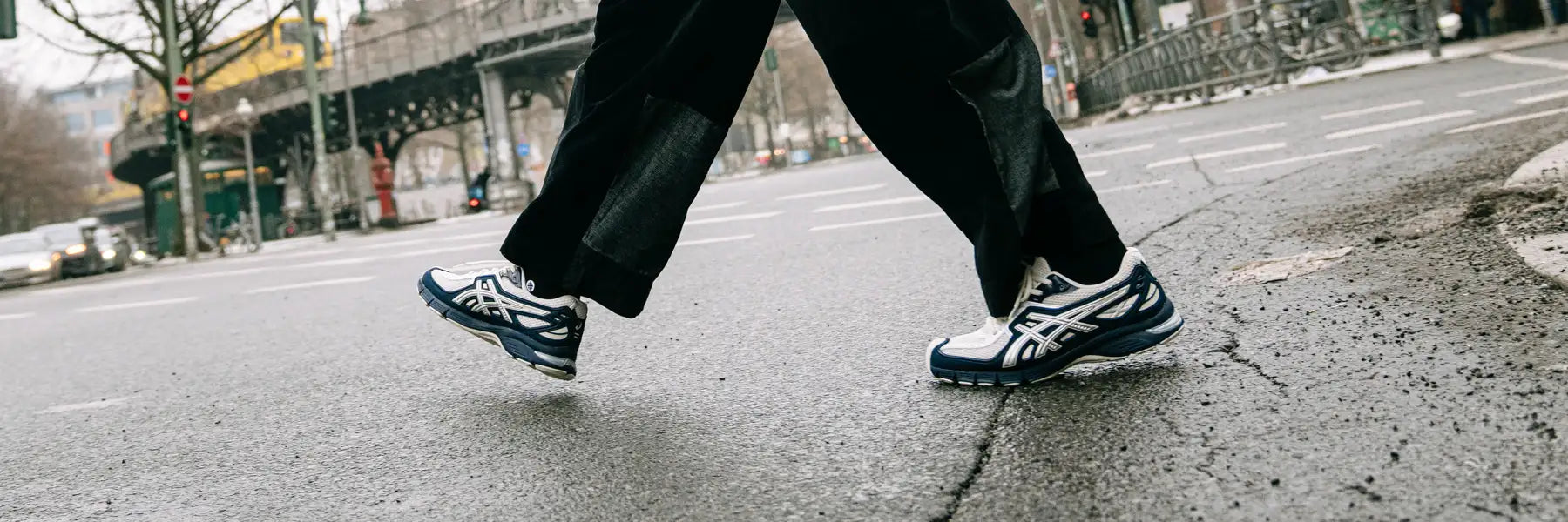 Person wearing black pants and blue and white Asics Gel-SD-Lyte Sneakers with blue accents crossing a wet city street, conveying a dynamic and urban atmosphere.