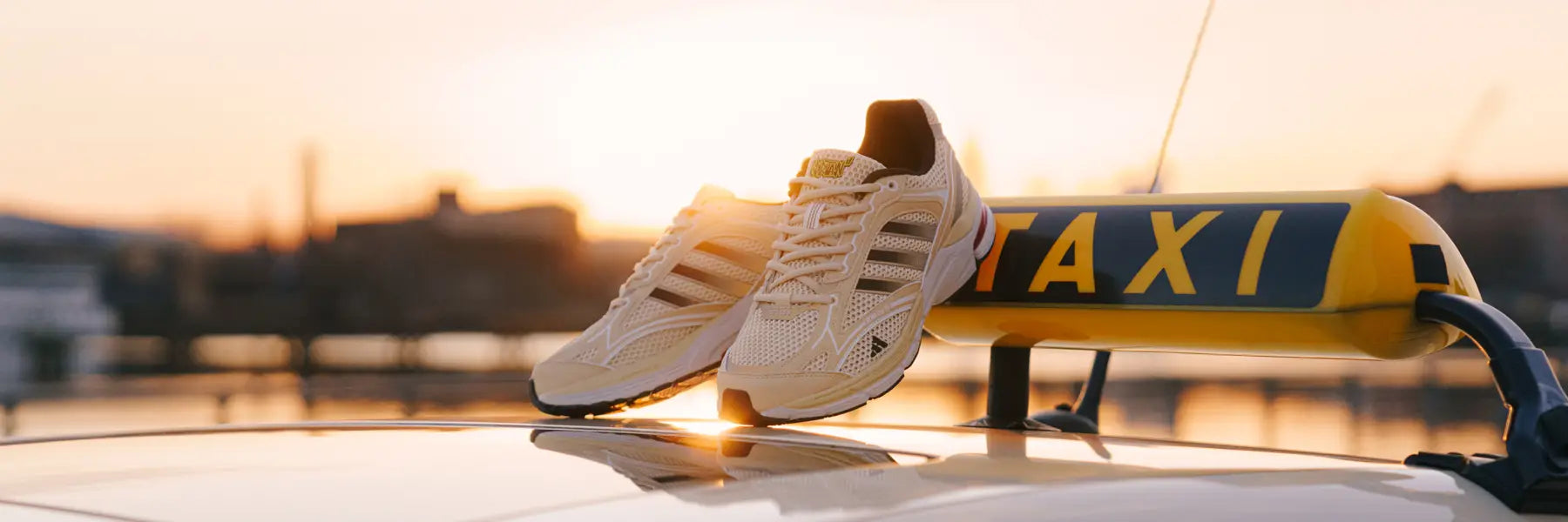 A pair of beige sneakers sit atop a taxi roof at sunset. adidas Spiritain 2000 Overkill Taxi. The warm glow casts a soft light, highlighting city buildings in the blurred background.