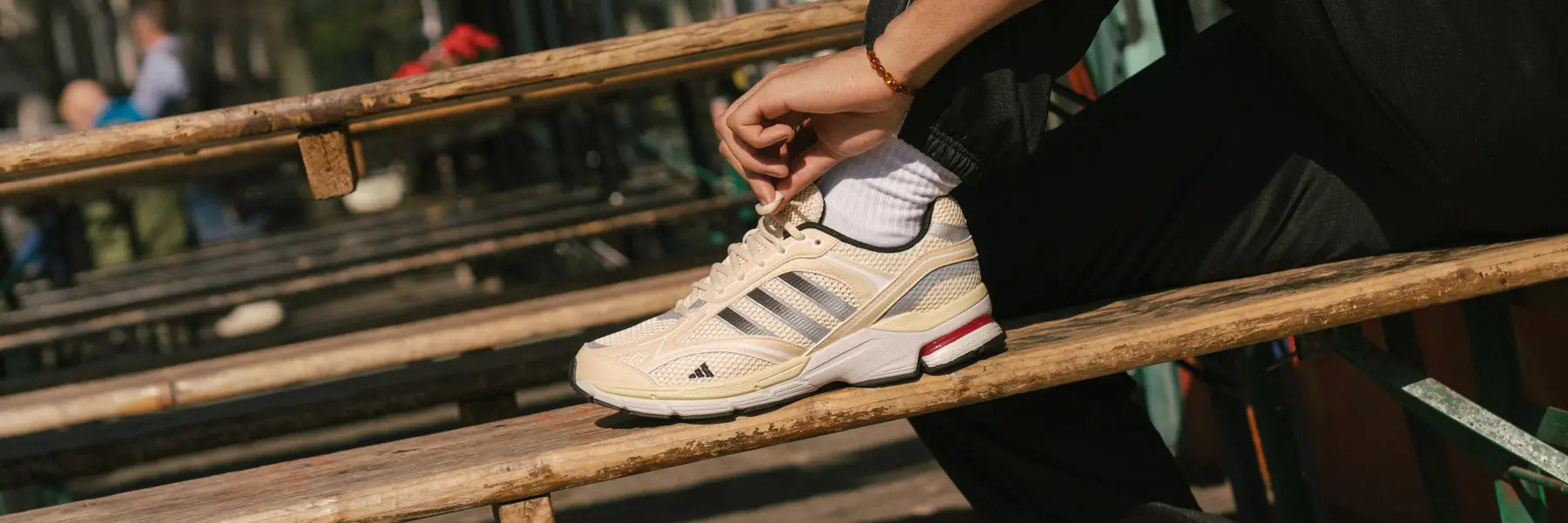A person in black pants adjusts their beige Adidas sneaker while sitting on wooden bleachers outdoors.