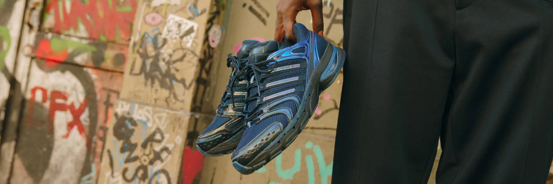 Hand holding a pair of blue and black athletic shoes like adidas Adistar Y2k sneaker with laces against a graffiti-covered wall