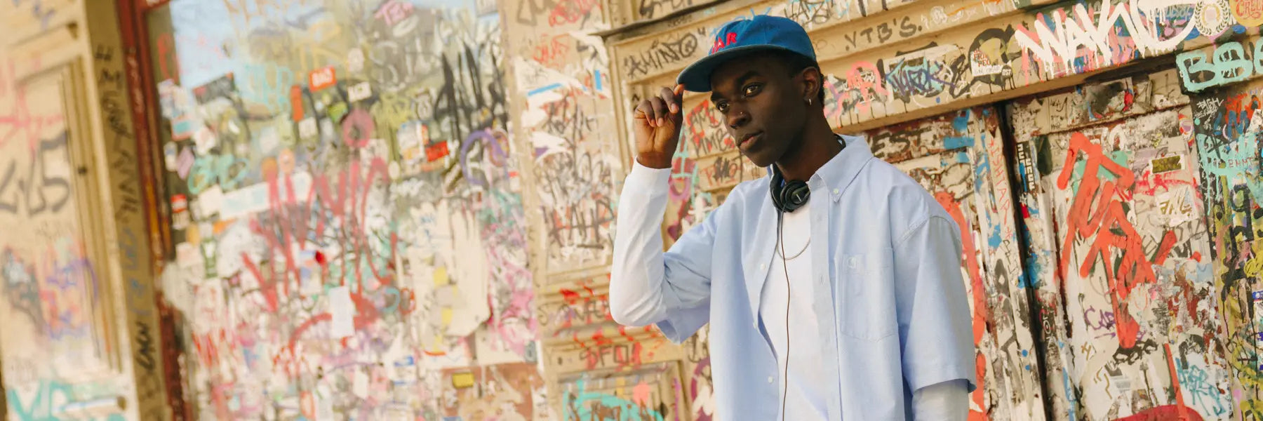 Young man in a blue cap and shirt stands in front of a vibrant, graffiti-covered wall, exuding a relaxed, urban vibe with headphones around his neck.